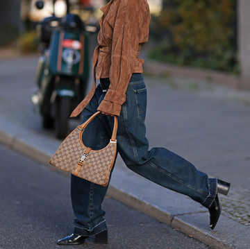 individual walking on a city street wearing a brown suede jacket and oversized blue jeans carrying a designer handbag