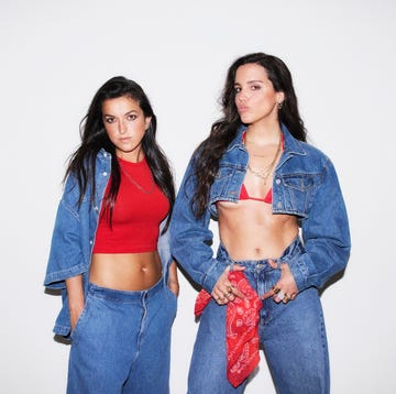 two women stand side by side against a plain white background one woman wears a red crop top with a denim jacket and high waisted denim shorts, while the other wears a red bikini top under a denim jacket and blue jeans they both have long, dark hair and accessorize with jewelry the woman on the right has a red bandana tied around her waist