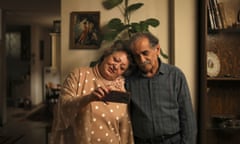 An elderly Iranian man and woman standing next to one another, her head on his right shoulder, look at a mobile phone she is holding in her right hand.