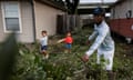 A man cleans up tree debris from a yard outside a home as his two children stand by