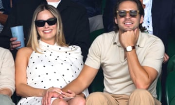 Margot Robbie seated in Centre Court wearing a white dress with black spots, alongside her husband. Both are wearing sunglasses