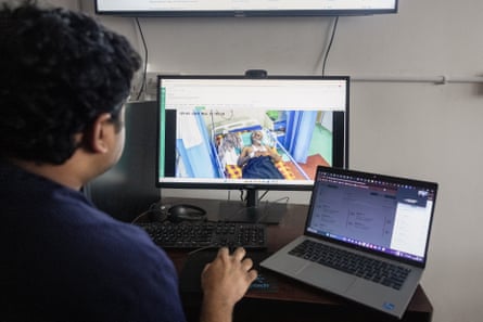 An Indian man seen from behind as he looks at a computer screen showing an older man lying on a hospital bed with a laptop next to it