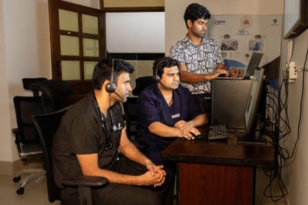 Two young Indian men wearing headphones sit at a desk looking at a screen while another man stands looking at a laptop