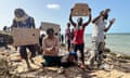 A small group of people on rocky ground next to a body of water holding placards, one reads 'Tunisia government is killing us slowly, we need help'