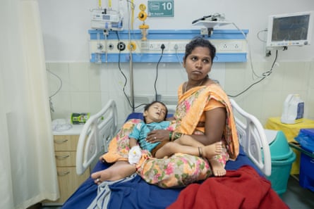 An Indian woman sitting on a hospital bed looks off camera as a small child with a canula in his hand lies on her lap