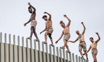 Five men in beige shorts balance with their arms out as they walk along a high narrow ledge