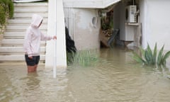 A person stands at the bottom of some steps, next to a house with flood water coming to above their knees