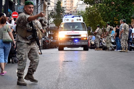 A soldier with a machine gun stands in a street in Beirut, pointing, as an ambulance passes a group of soldiers behind him