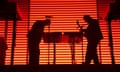 Two male musicians, standing next to laptops on tables and microphones on stands, in silhouette against an illuminated orange striped background.