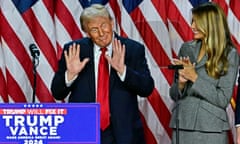 Donald Trump smirks on stage with his palms in the air as Melania Trump looks on, smiling and clapping. Several US flags are draped behind them