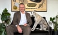 Benjamen Judd in a brown tweed suit and white shirt, sitting on a couch with dog beside him