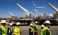 workers wearing neon vests and white hardhats walk by wind turbines under construction