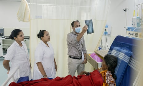 An Indian doctor stands by a patient in a bed holding up an X-ray while two female medics in white coats stand beside him