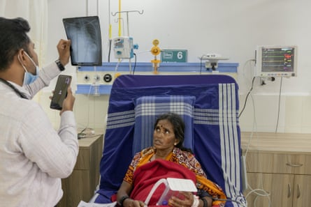 An Indian man holds up an X-ray while talking into a phone as an anxious-looking older woman looks at him from a hospital bed 