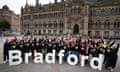 Volunteers celebrate the launch of Bradford 2025 UK City of Culture: they are lined up and cheering behind large white letters spelling out the city's name, and wear black T-shirts with a B shaped to look like a heart. They are standing in a large paved plaza in front of a large, historic stone building