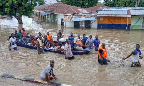 A group of about 20 people on a boat, with several others standing in the deep water. A cluster of buildings, half submerged in water, can be seen behind them.