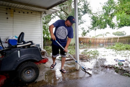 A man wearing a blue T-shirt and shorts sweeps away water outside a home