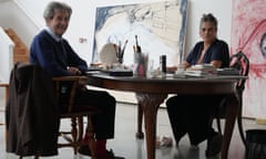 Melvyn Bragg sitting at a table opposite Tracey Emin in her studio with her paintings in the background.