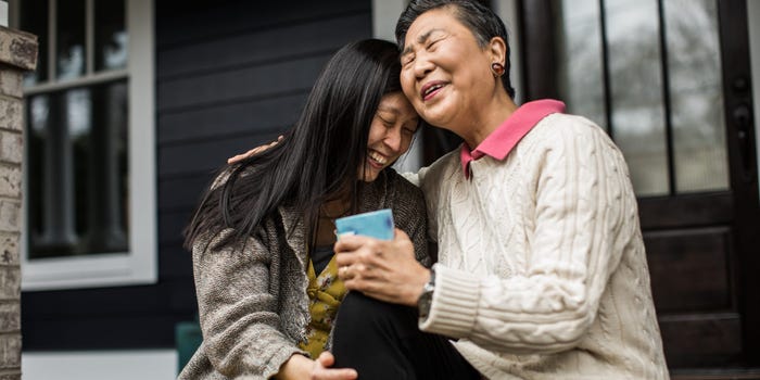 An older woman and her 20-something daughter sit on the steps of their front porch, hugging