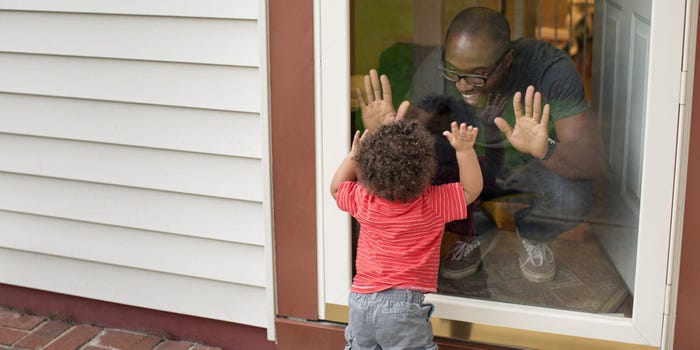 A dad in a home with a second mortgage makes a silly face at his toddler through a glass-screen front door.