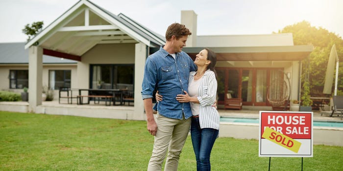 Couple stands in front of home, covering for sale sign with a sold sign