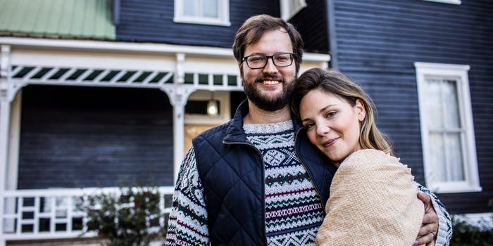 30-something-year-old man and woman embrace in front of a dark blue, two-story house