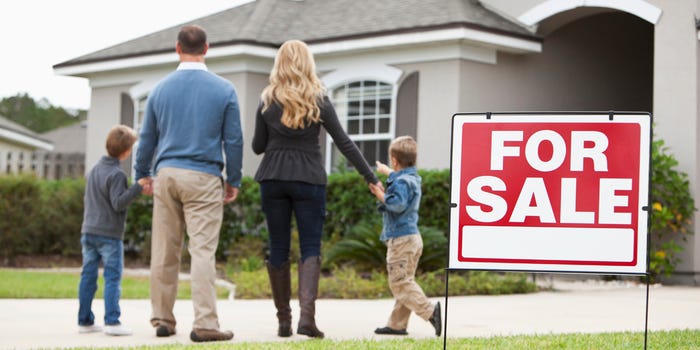 A photo of a young family in front of a house with a for sale sign in the foreground.