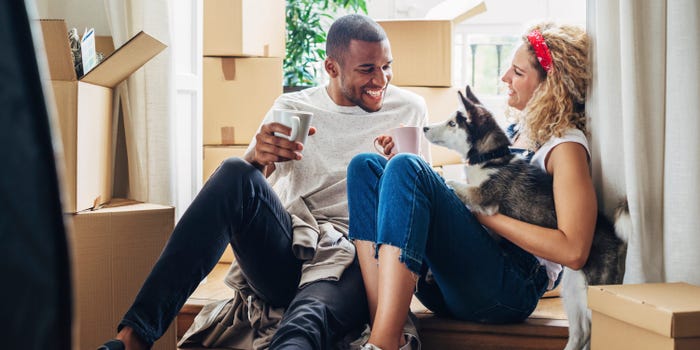 A man and woman who debated renting vs. buying sit in the doorway to a house surrounded by boxes with their dog.