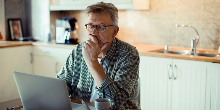 A middle-aged man sits in his kitchen and looks at his laptop