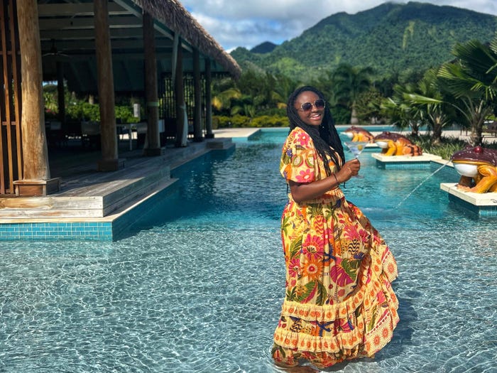 Mariette stands in a shallow pool in a long, patterned dress. On her left is a hut-like building, and on her right are frog statues that shoot water. Behind her are trees and greenery-covered mountains.