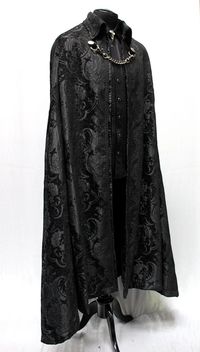 "A magnificent cape fit for royalty. Made in rich flowing black tapestry fabric with rich black satin lining inside. The high pointed collar can be worn down or up for an extra dramatic effect. Braided black trim lines the collar and along the front of cape. Fully lined in rich black satin. Fastens in front with a heavy antiqued chain which clasp at either side to heavy D-rings and decorated with two ornate metal buttons. Slits in the front of cape allow the wearer to reach through and hold a be