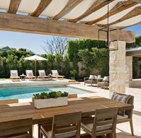 Stone pillars accent a rustic wood pergola finished with canvas and positioned over a teak plank dining table surrounded by teak slatted back dining chairs placed in front of an in ground swimming pool.