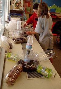 Two students work on their biodome. Nearby are unique model biodome designs, all made with conjoined clear plastic bottles and trays.