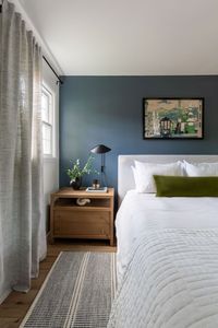 Natural light. Blue walls. Bedside table. Black table lamp. Throw pillows. White bedspread. Striped throw rug. Scandinavian