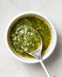 A spoon dipped into a bowl of compound butter with herbs and garlic on a white surface.