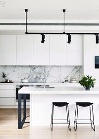 Black and white monochrome kitchen: handleless white cabinets and benchtops, grey marble splashback, black bar stools, black spotlights on suspended ceiling track, timber floorboards