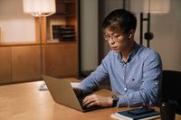 Focused asian guy in earphones working with laptop while sitting at table in office