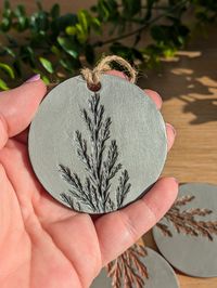 Handmade Iridescent Graphite Clay Ornament - Winter Tree Design Add a touch of elegance to your holiday décor with this stunning handmade clay ornament. Crafted in Wales, each piece features a beautiful conifer tree design, painted in iridescent graphite acrylic and sealed with a glossy finish for protection and shine. Perfect as a Christmas decoration or a thoughtful gift! Customization Options: Choose your desired quantity and twine colour: ✧ Twine Colours: Natural Twine White Cotton Green Bak