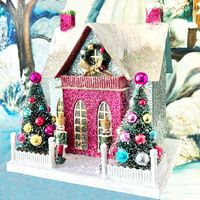 Festive blue glitter Chalet with 2 sparkling red chimneys, a hot pink front, pink balcony, two ornament encrusted bottle brush trees and a pair of Nutcrackers standing guard. 12" Tall x 10.5" Wide x 8" Deep