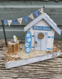 A wooden weathered beach shack with a handcrafted wooden surfboard and a clay seagull. This glorious little beach hut has all the charm of the full sized ones on local beaches. The weathered look and pastel tones would make the perfect addition to any beach themed home. With a hand-painted surfboard, this scene would make the perfect gift for surfers. A pesky seagull keeps and eye out for tasty treats. Made from reclaimed wood and deckchair wood, this is finished with beach sand, shingle and paper ticket bunting made from original deckchair tickets.