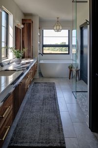 bathroom chandelier, stand alone tub, glass and tile shower, floor rug runner, doubel sinks, double mirrors, natural stone black and white counter tops, tile floor, stained wood cabinets, walnut wood cabinets, brass hardware