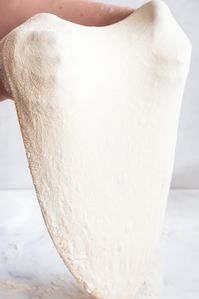 pizza dough being stretched on the back of two hands in mid air. the dough stretches down to the counter, just brushing against it.