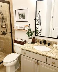 Light wood and bronze shelf is mounted above a white toilet standing next to a beige washstand. A rectangle black bathroom mirror is hung above the beige and brown granite countertop fitted with a black faucet kit.