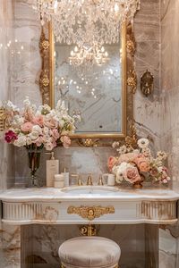 Step into luxury with this elegantly designed bathroom vanity area featuring a white marble sink, ornate gold mirror, and sparkling crystal chandelier. The opulent space is adorned with lush floral arrangements, gold fixtures, and a plush, vintage stool. Experience the sophisticated and classic ambiance with hints of a bygone era's aesthetic. #LuxuryBathroom #ElegantDesign #ClassicAesthetic