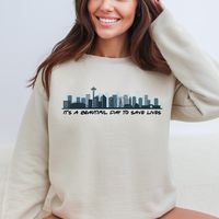 Introducing our Grey's Anatomy It's A Beautiful Day To Save Lives Sweatshirt, a must-have for die-hard fans and the perfect gift for moms who love the show! Crafted with comfort in mind, this sweatshirt features a captivating graphic showcasing the Seattle Skyline. Indulge in cozy warmth while binge-watching your favorite medical drama or gift this stylish piece to a Grey's-loving mom who deserves the best. Elevate your fandom and stay snug with this iconic sweatshirt that speaks volumes to Grey