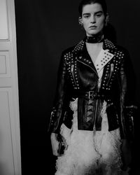 A black biker jacket is finished with matt and shine studs, a painted lapel and quilted panelling. Worn with an exploded asymmetric knit skirt with ruffles in a fine silk rib. Photographed backstage at the Alexander McQueen Autumn/Winter 2019 show.