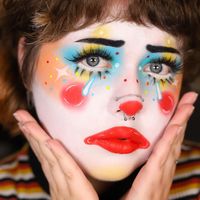 This look radiates joy and whimsy with its vibrant color palette. The artist has used a white base and adorned the face with multicolored accents, including blue eyeshadow, yellow blush, and red lips. The dramatic eyelashes and dotted patterns on the cheeks and around the eyes add a playful, colorful touch. The red clown nose and subtle glitter create a cute yet simple appearance, making this look both eye-catching and approachable. This inspiration is perfect for anyone looking to bring a burst of color and creativity to their clown makeup repertoire.   Photo credit by: @katie.artistry