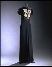 Elsa Schiaparelli Haute Couture evening dress from 1940, a short sleeved dress in matt black crepe.The extremely simple, fluid shape is relieved by a slashed neckline and a bold embroidered spray of lilies by the House of Lesage in various sizes of pearls, bead, sequins and metallic strip. The dress fastens with a black plastic zip on the left-hand side. House of Schiaparelli.