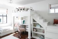 A pink velvet chair sits at a white desk with a chrome trim beneath a white loft bed finished with a white staircase located over a white built-in bookcase.