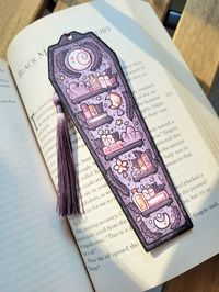 "2\"x6\" Purple Coffin Bookmark with Tassel (Not Laminated): This spooky cute handmade coffin shaped bookmark features the illustration of a variety of witch bookshelves adorned with various colored stacked books, hanging star and moon, candles, potions and crystals decorated with floral/plant motifs and moon designs on a purple coffin background with a purple tassel! Made of cardstock paper, it is the perfect autumn/fall book marker accessory for book lovers to use in your favorite book, journa